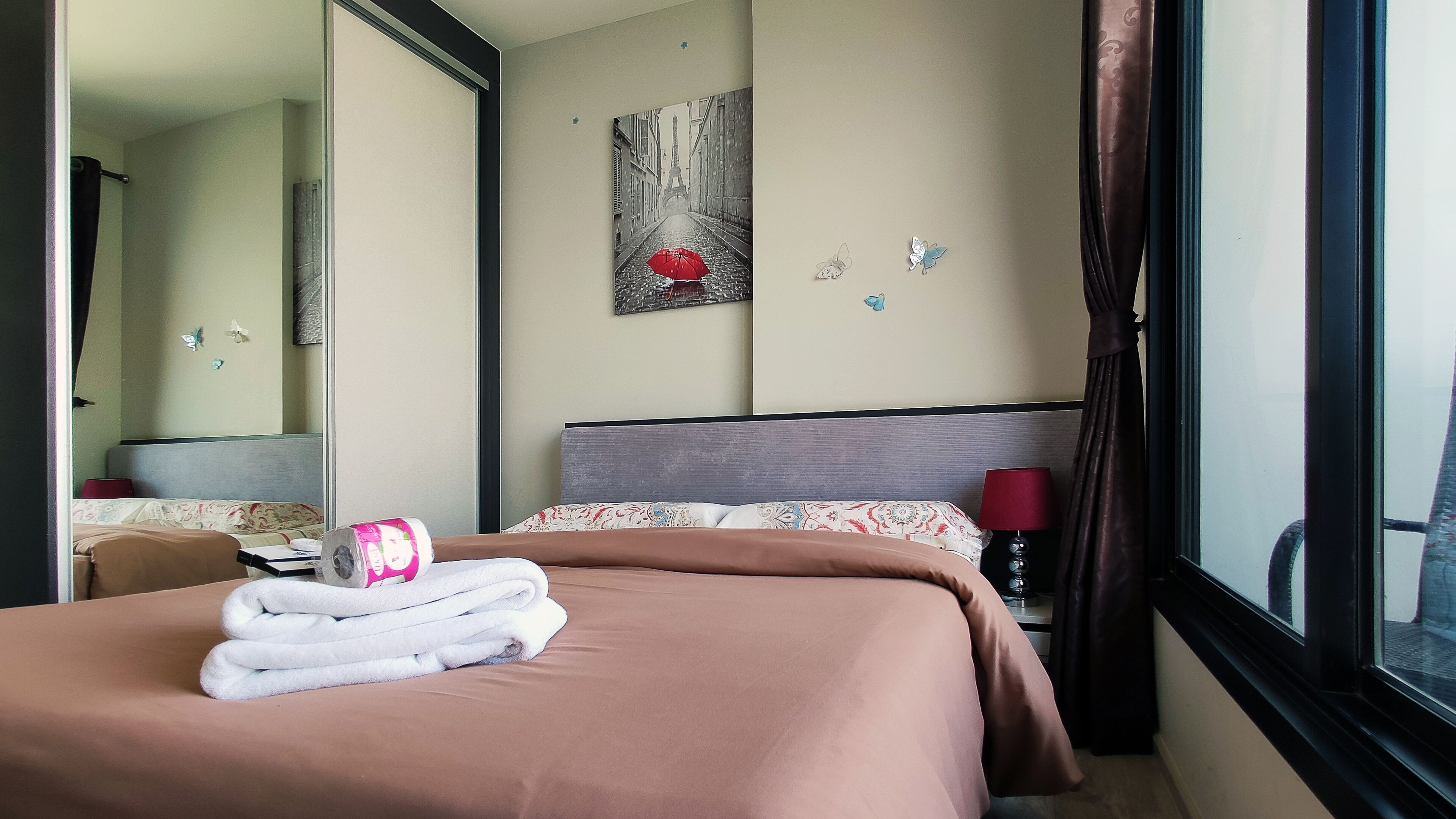 A hotel room with a bed, a bedside table, a lamp, a mirror, and a window with curtains.