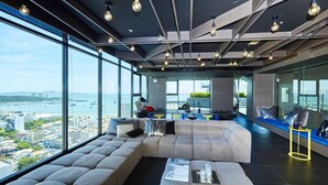 A modern lounge area with a large sectional sofa, a coffee table, and a view of the city and water. - Pattaya Central Sea View Pool Suite (Pattaya)