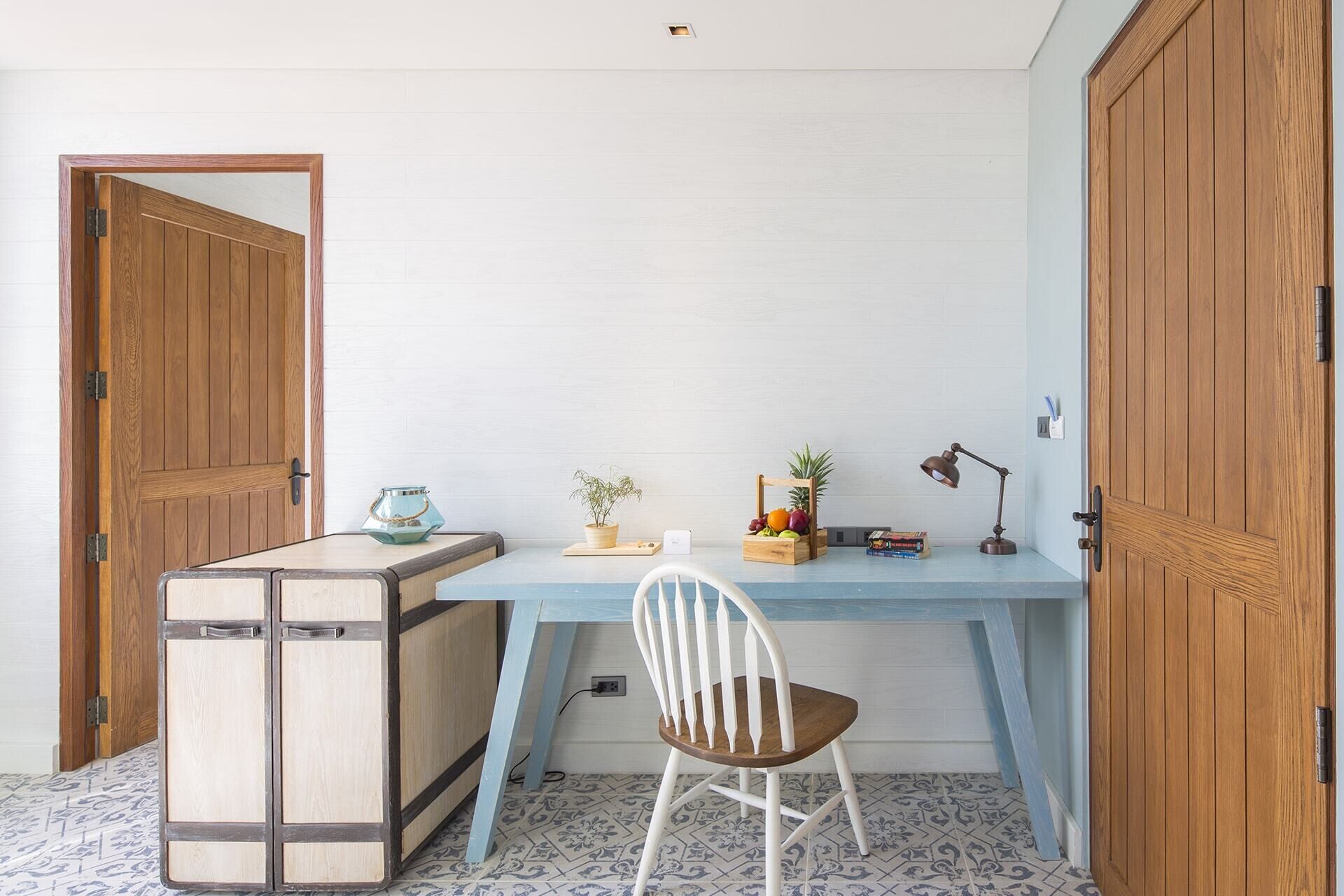 Una habitación con un escritorio de madera, una silla blanca, una planta pequeña, una lámpara y un armario de madera.