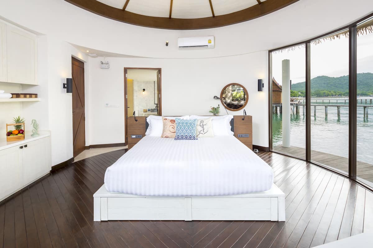 Un dormitorio moderno con una cama grande, suelo de madera, ventilador de techo, vista a un cuerpo de agua y una puerta de madera.