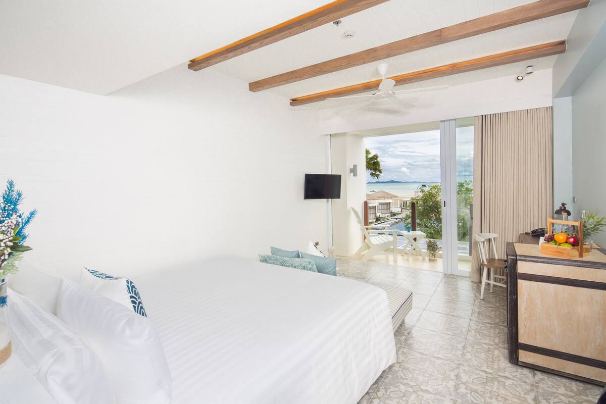 Habitación de hotel con cama, escritorio con frutero, televisión y vista al océano.