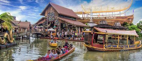 Un marché flottant avec des bateaux traditionnels en bois, un grand bâtiment en bois portant l’inscription « Marché flottant de Pattaya » et un grand navire en arrière-plan.