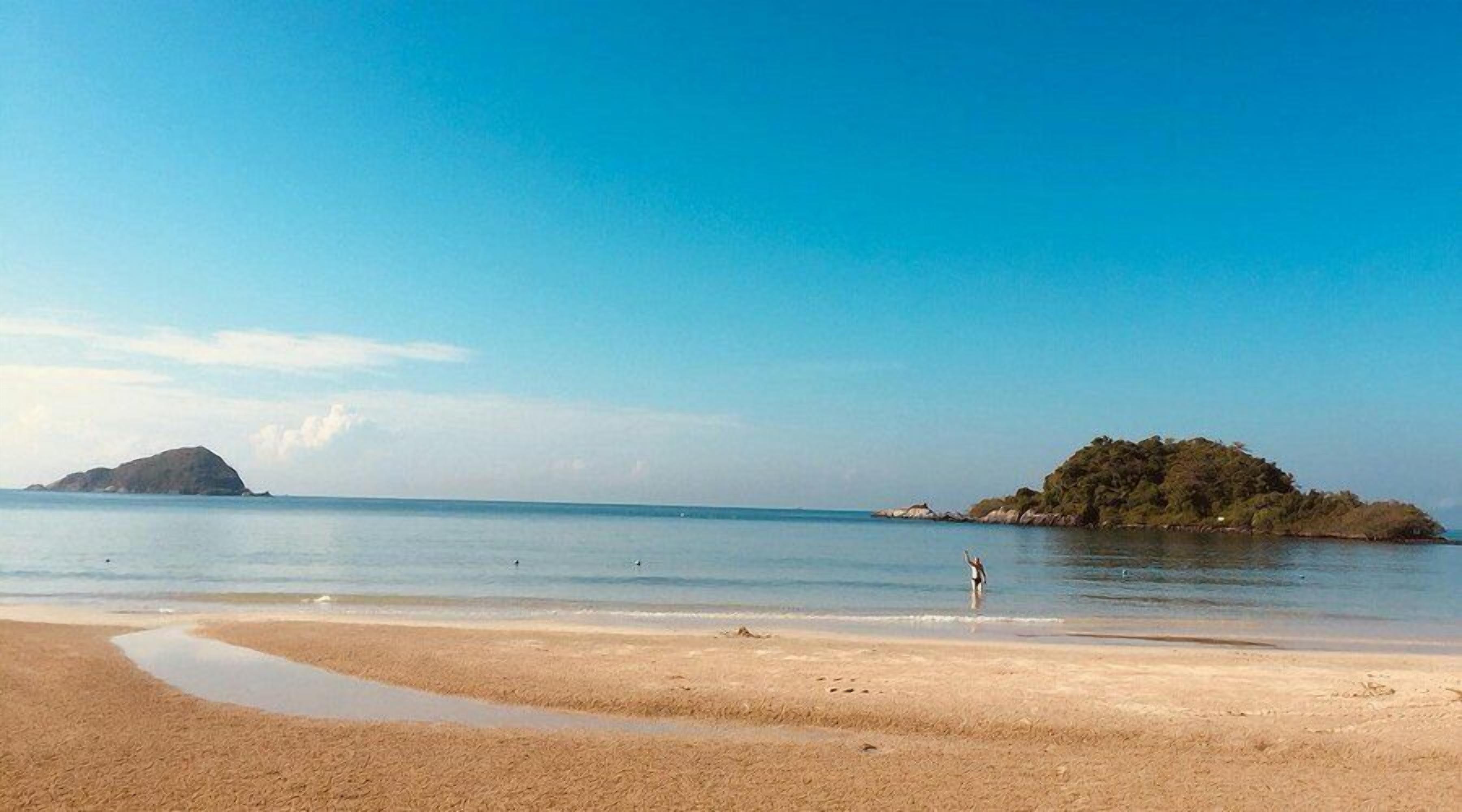 A beach with a clear sky, calm sea, and a small island in the distance.