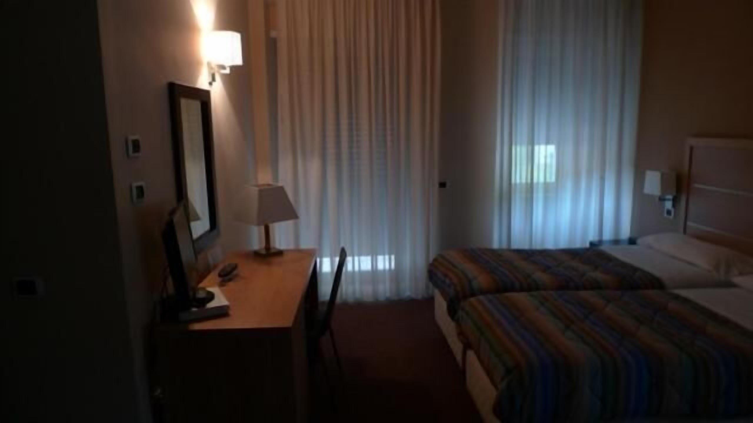 Quarto de hotel com cama, escrivaninha com cadeira, luminária e espelho.