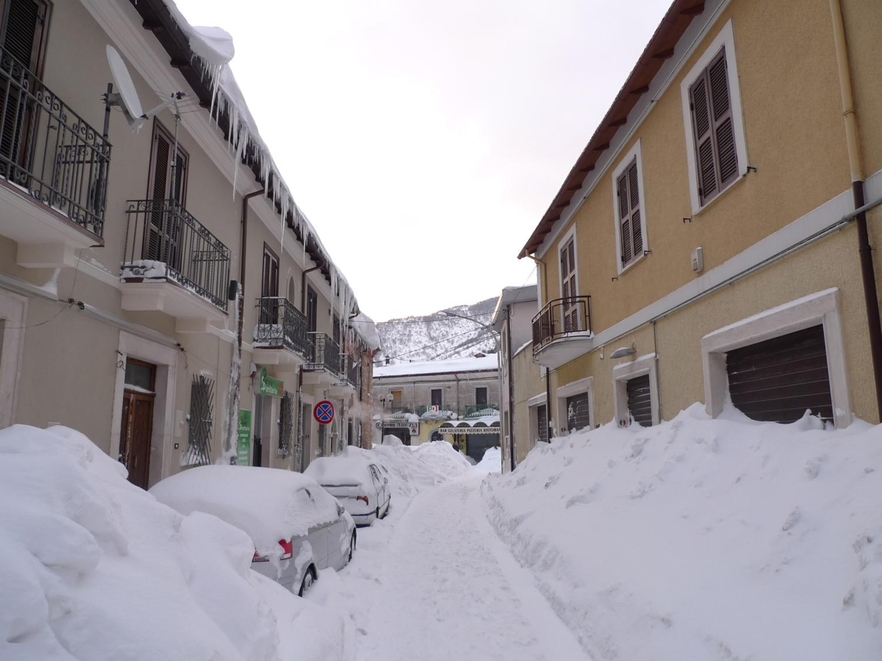 A narrow street with snow-covered cars and buildings, featuring balconies and a mountain in the background.
