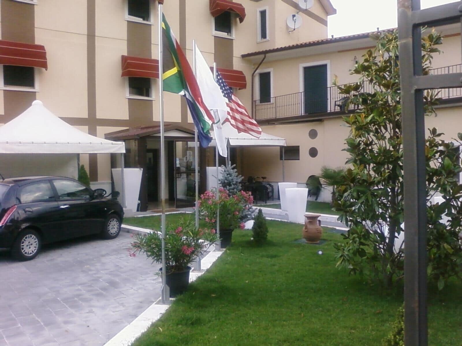 A hotel entrance with flags, a car, and a garden area.