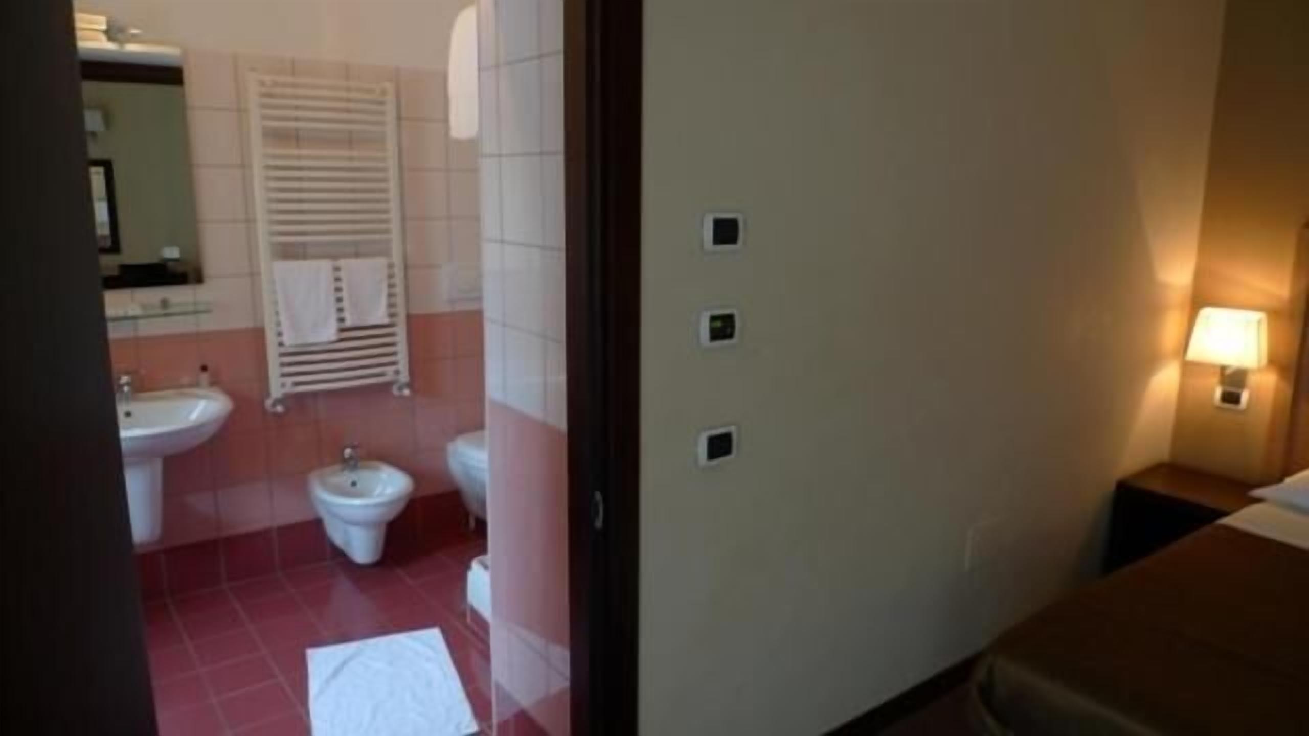 Un bagno d'albergo con lavandino, bidet e WC. C'è un asciugamano sul pavimento e un comodino con una lampada.