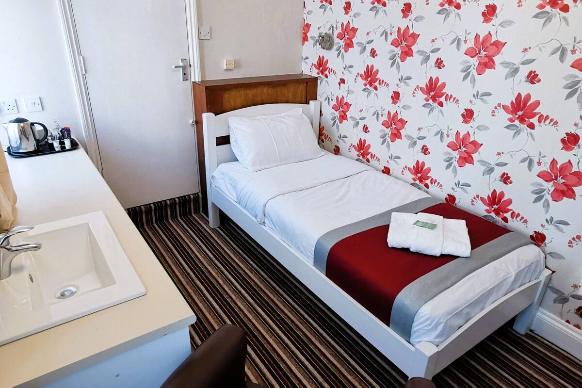 A hotel room with a single bed, a bedside table with a lamp, a small sink, and a floral wallpaper.
