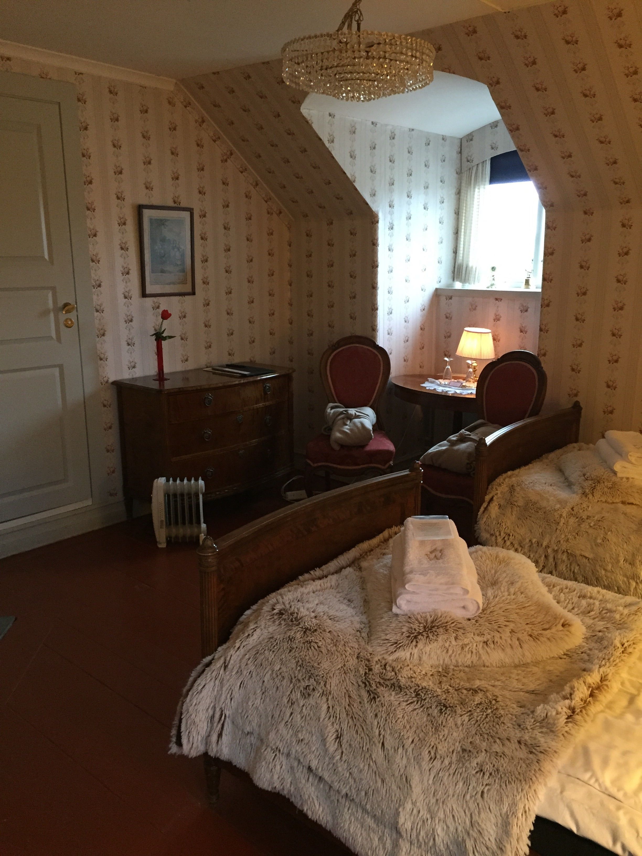 Een slaapkamer met een schuine balkenlaag, een houten bed met een zachte deken, een houten dressoir, een stoel en een bureau met een lamp.
