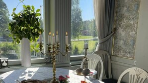 Une table à manger dressée avec une variété de plats, notamment des fruits, du pain et des boissons, dans une pièce dotée d’une grande fenêtre et d’une plante en pot.