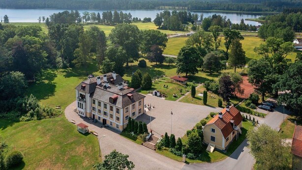 Un grand bâtiment à plusieurs étages, doté d’une cour intérieure, entouré d’une végétation luxuriante et avec un lac en arrière-plan.