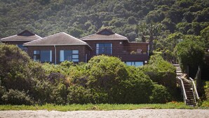 A beachfront property with a wooden staircase leading to a balcony, surrounded by lush greenery and a sandy beach. - ArchRock Resort (Keurboomstrand)