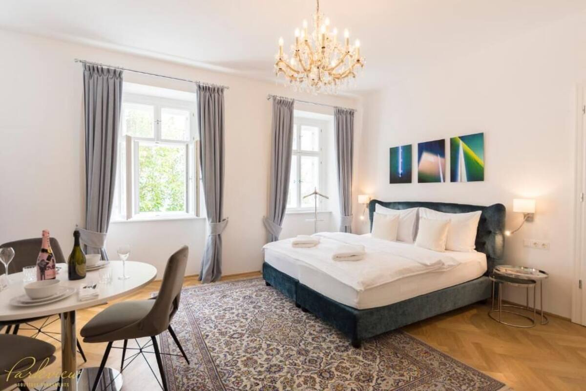 A hotel room with a large bed, a chandelier, a dining area with a table and chairs, and artwork on the wall.