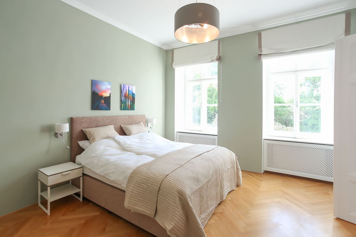 A bedroom with a bed, bedside table, two wall-mounted lamps, and two windows with blinds.