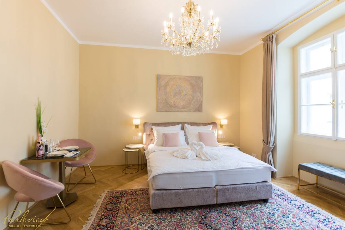 A hotel room with a large bed, a chandelier, a desk with a chair, and a window with curtains.