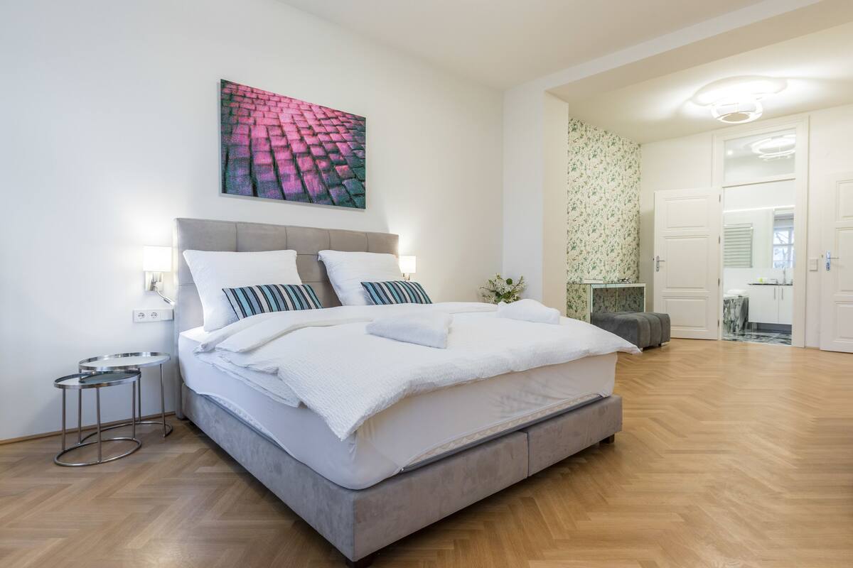 A modern bedroom with a large bed, a headboard, bedside tables, and a wall-mounted artwork.