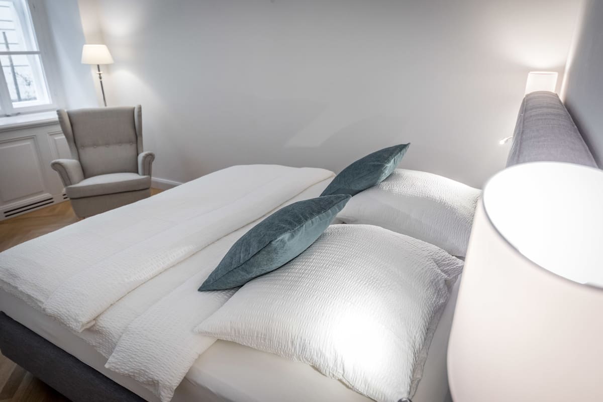 A neatly made bed with white linens and a single teal pillow, a grey armchair, and a bedside lamp.