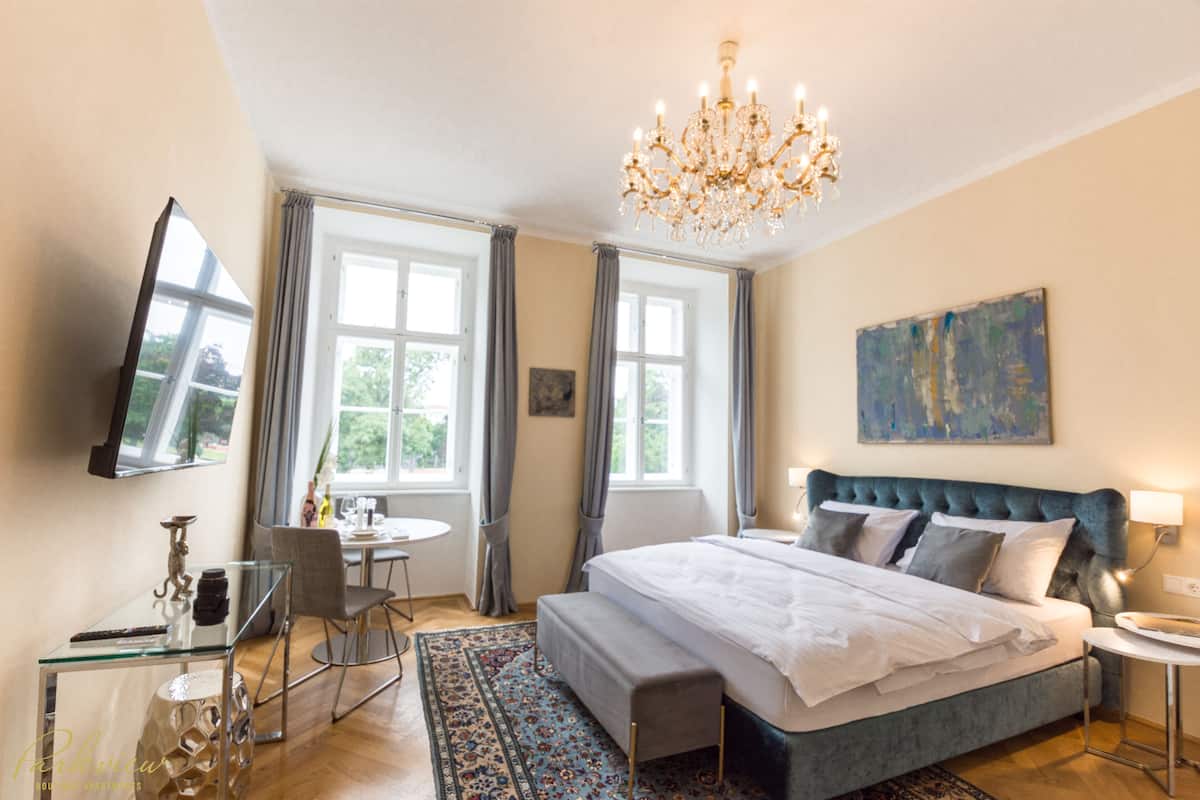 A hotel room with a large bed, a chandelier, a flat-screen TV, a dining table, and a window with curtains.