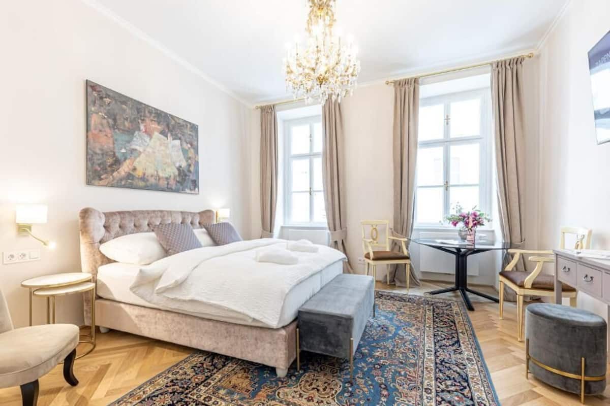 A well-lit bedroom with a large bed, a chandelier, a painting on the wall, and a view of the outside through the window.