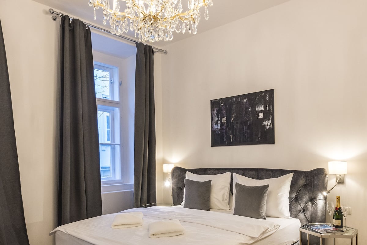 A modern bedroom with a large bed, a chandelier, a nightstand with a bottle of champagne, and a patterned rug.