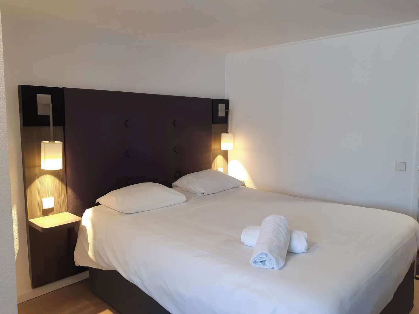 Een hotelkamer met een groot bed, twee bedlampen en een opgemaakt bed met een opgerolde handdoek.