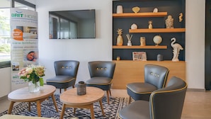 A hotel reception area with a modern design, featuring a flat-screen TV, comfortable seating, and decorative shelves with various items. - Brit Hotel Reims la Neuvillette (Reims)