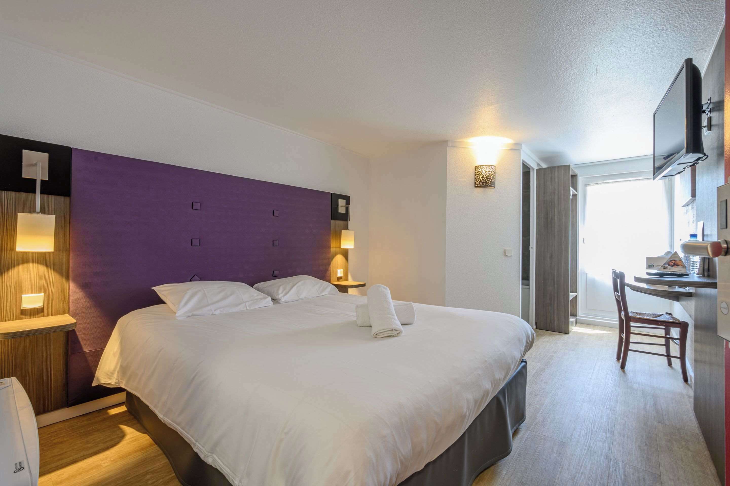 A hotel room with a large bed, a desk with a chair, a television, and a window with curtains.
