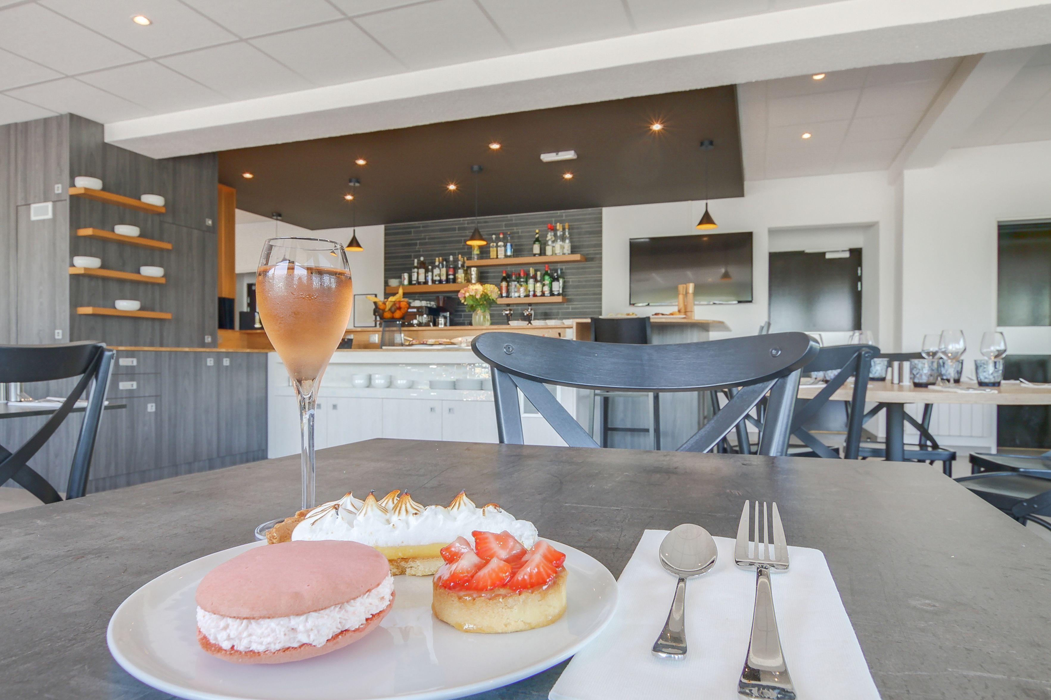 A dining area with a table set for two, featuring a dessert plate with a pink macaron and a strawberry-topped pastry, a glass of a light-colored beverage, and a bar area in the background.
