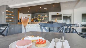 A dining area with a table set for two, featuring a dessert plate with a pink macaron and a strawberry-topped pastry, a glass of a light-colored beverage, and a bar area in the background.