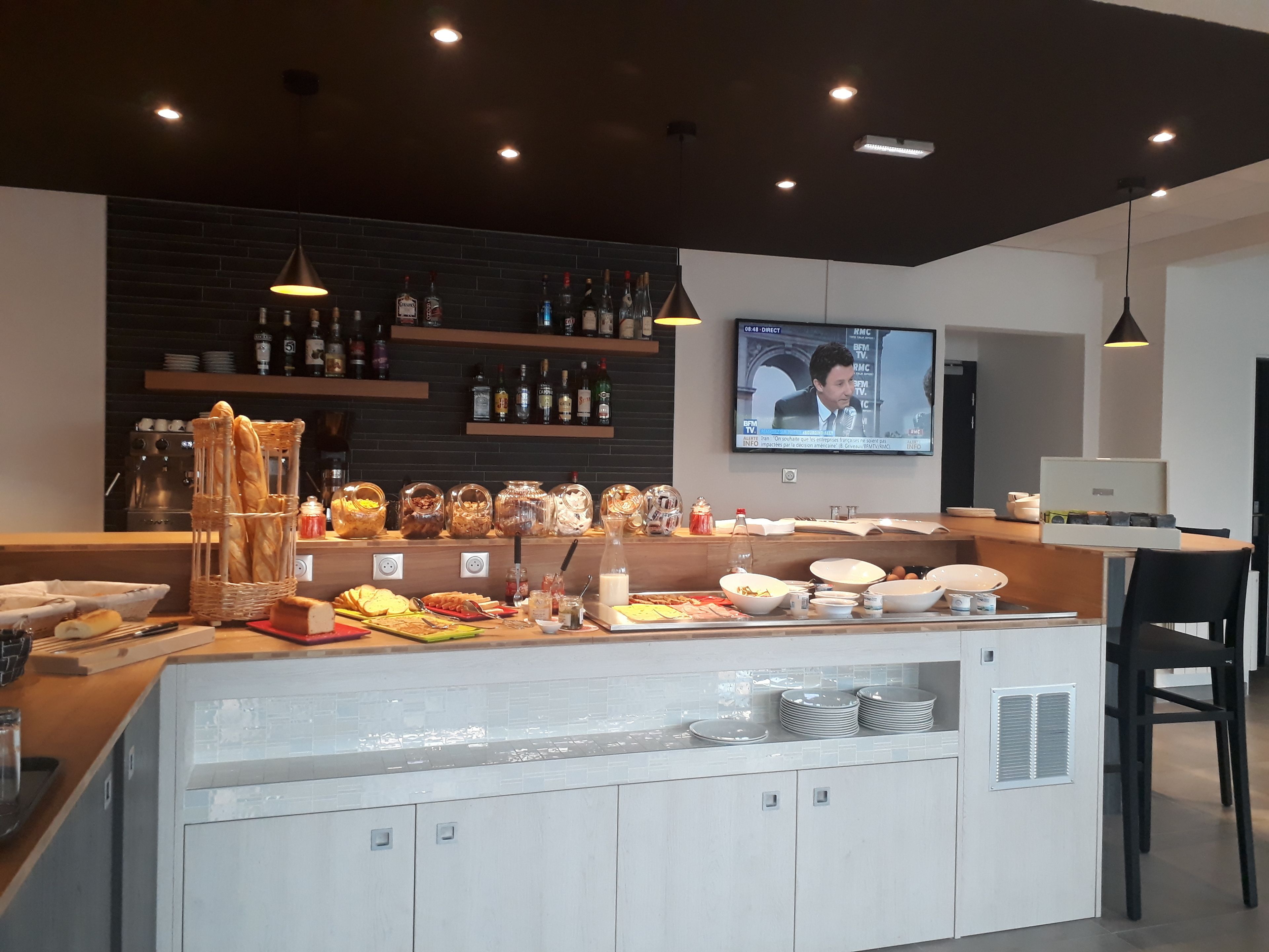 A modern bar area with a television displaying a news program, a variety of food and drink dispensers, and a seating area with a laptop and a chair.