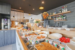 Un buffet proposant une variété de plats, notamment des œufs, du yaourt et des viennoiseries, installé dans un espace cuisine moderne agrémenté d’éléments en bois et de luminaires suspendus.