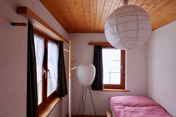 A bedroom with a wooden ceiling, a window with curtains, a bedside table, and a hanging paper lantern. - Ferienwohnung Sandgasse (Glarus Sued)