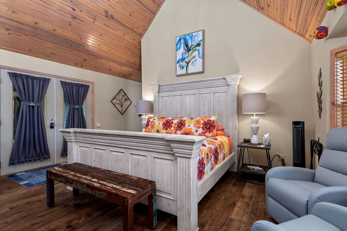 A bedroom with a wooden ceiling, a bed with floral bedding, a bedside table with a lamp, and a blue armchair.