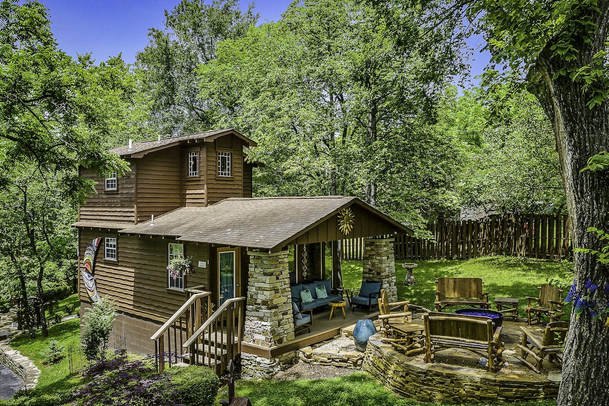A rustic cabin with a stone porch, wooden deck, and outdoor seating area surrounded by lush greenery.