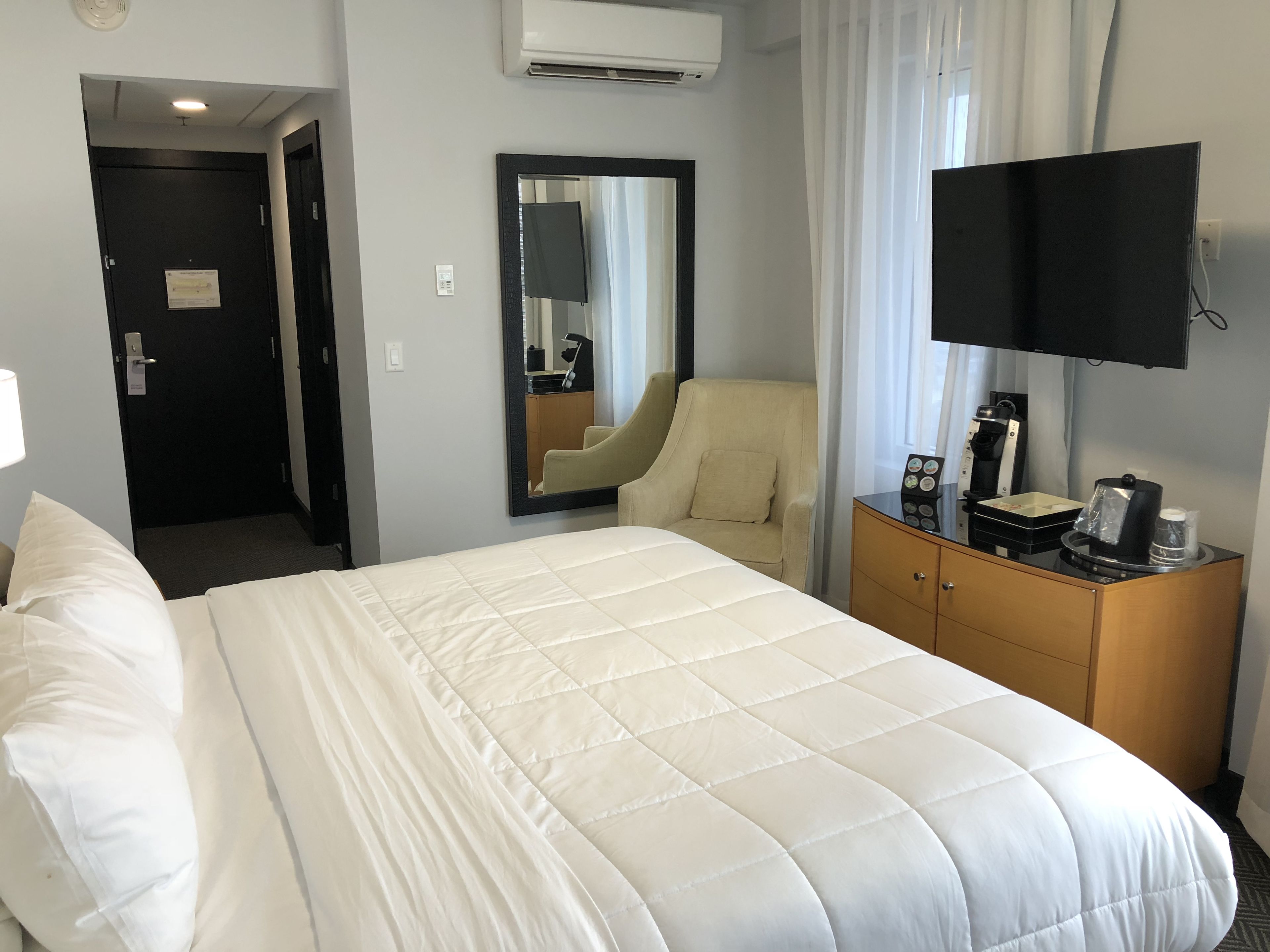 A hotel room with a large bed, a chair, a dresser, a flat-screen TV, and an air conditioning unit.