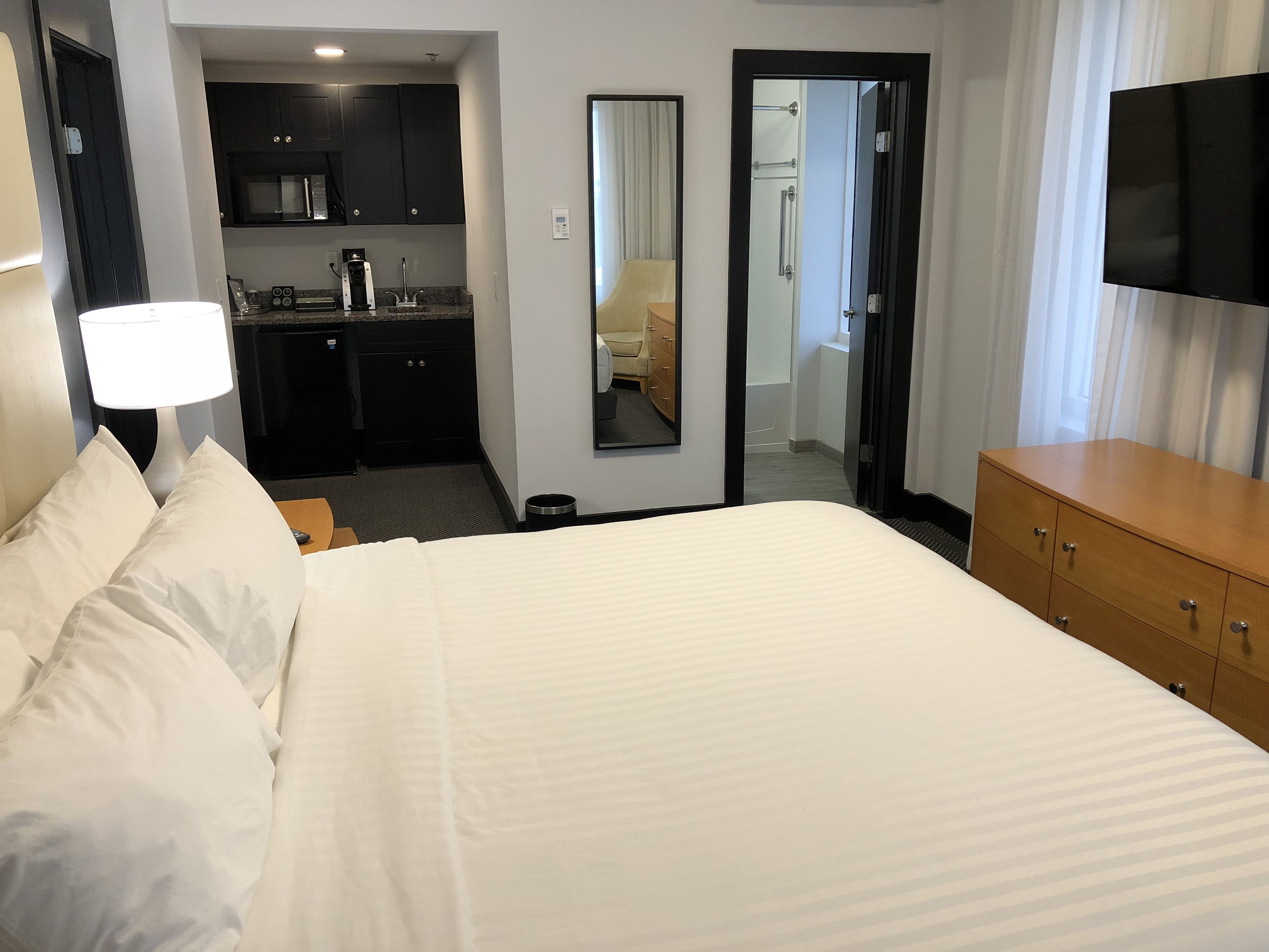 Et hotelværelse med en stor seng, en trækommode, et fladskærms-tv, et skrivebord med kaffemaskine og et tekøkken.