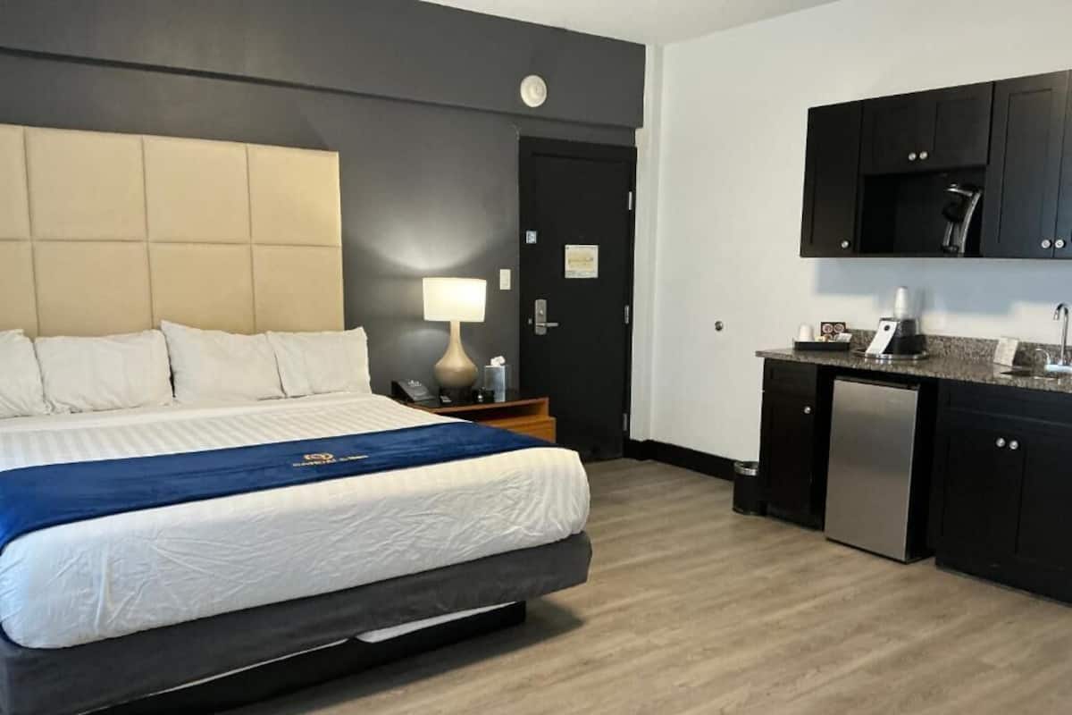 A hotel room with a bed, bedside table, lamp, and a kitchenette area with a sink and cabinets.