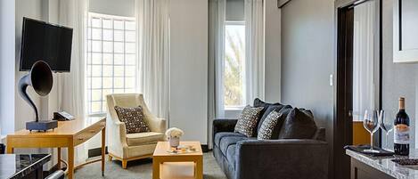A modern living room with a sofa, armchair, and coffee table. There is a flat-screen TV mounted on the wall, a wine bottle, and glasses on a counter.