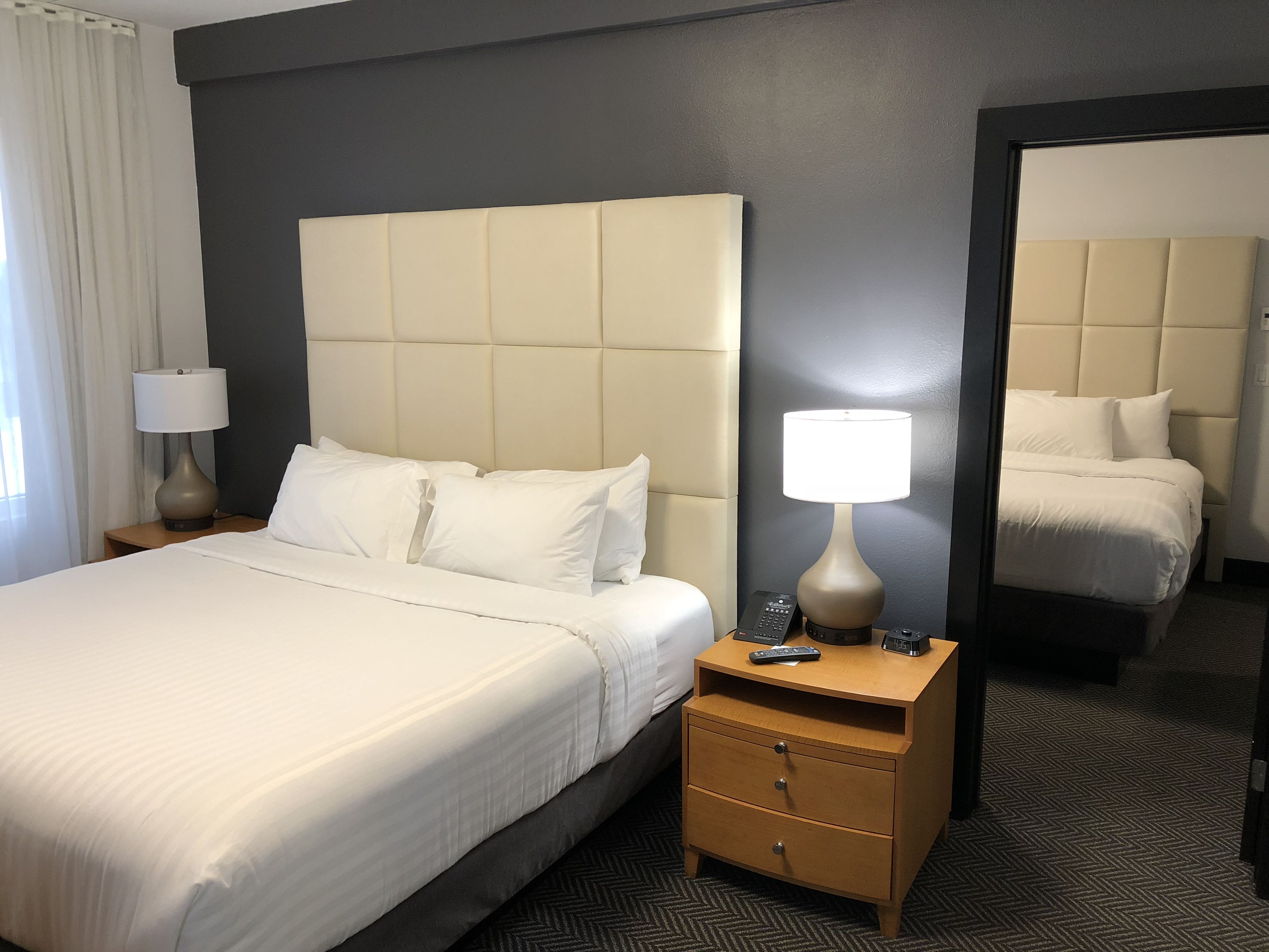 Et hotelværelse med en stor seng, et natbord med lampe, et spejl og en kommode.