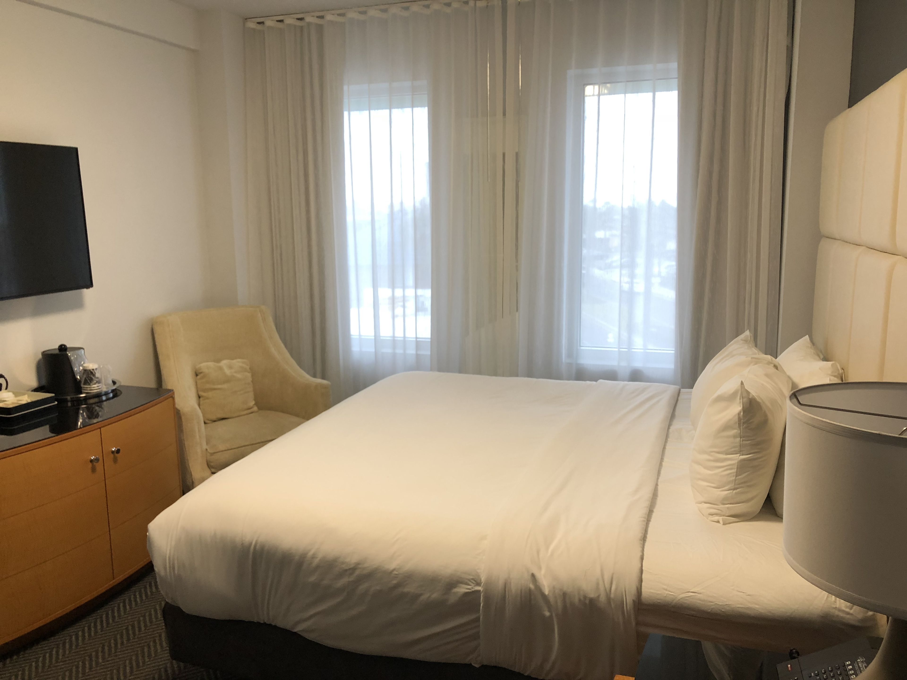 A hotel room with a bed, a chair, a TV, a desk with a coffee maker, and a window with curtains.