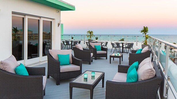 Une terrasse dotée de meubles en osier, de tables en verre et offrant une vue sur la mer au coucher du soleil.