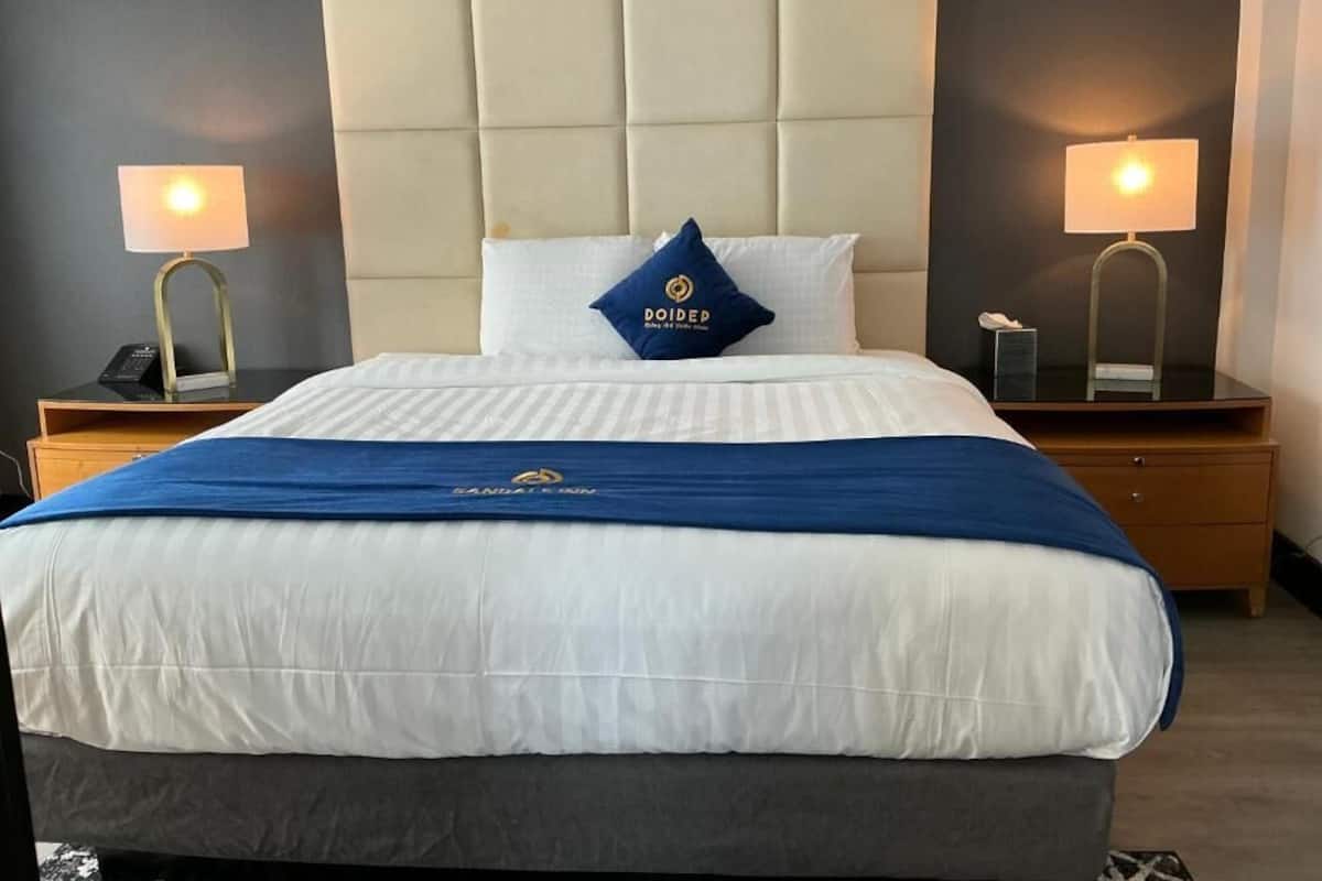 A hotel room with a large bed, a blue and white bedspread, a blue pillow with a logo, two bedside lamps, and a wooden nightstand.