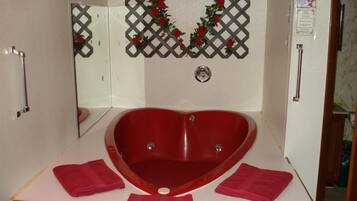 A heart-shaped bathtub in a bathroom with a mirror, a decorative lattice with roses, and two towels on the counter.