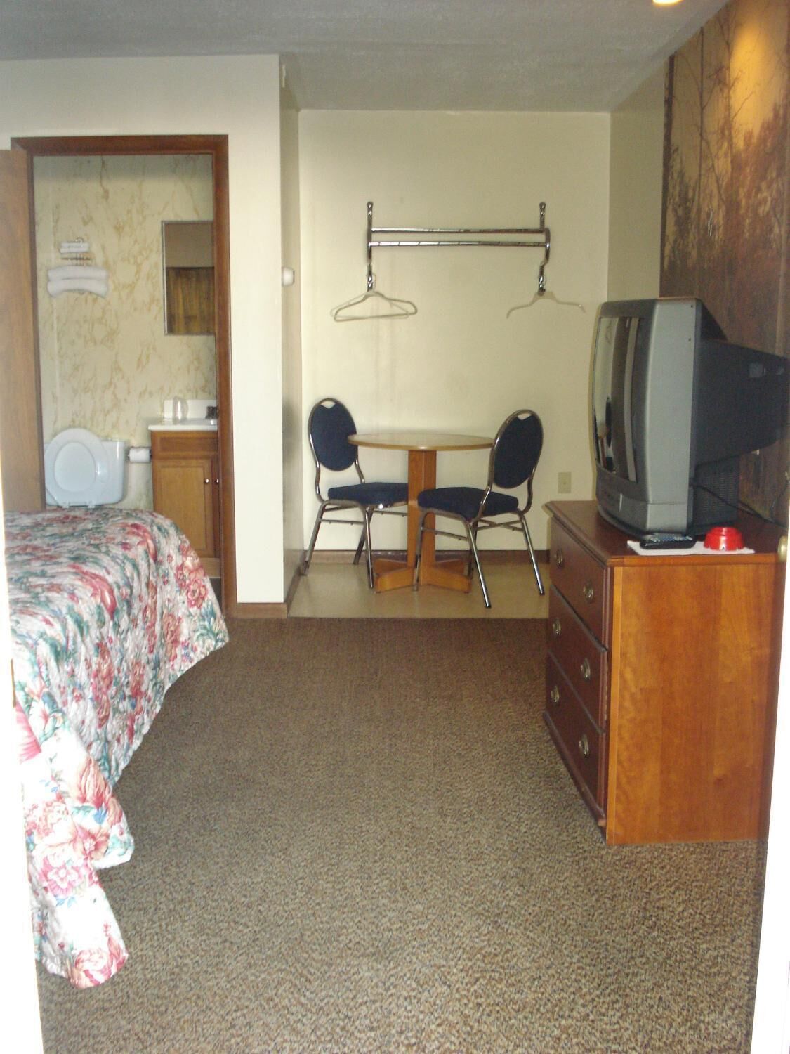 A small, compact room with a single bed, a wooden dresser, a television, and a small table with two chairs.