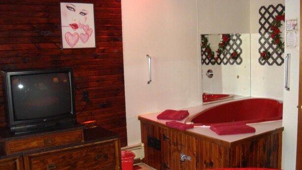A room with a wooden dresser, a television, a red carpet, a large mirror, and a red bath mat. - Cedar Grove Motel (Altoona)
