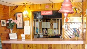 A reception desk with a 'Welcome' sign, a 'Vacancy' sign, and a bell above it. There are framed pictures and a vase with flowers on the desk. - Cedar Grove Motel (Altoona)