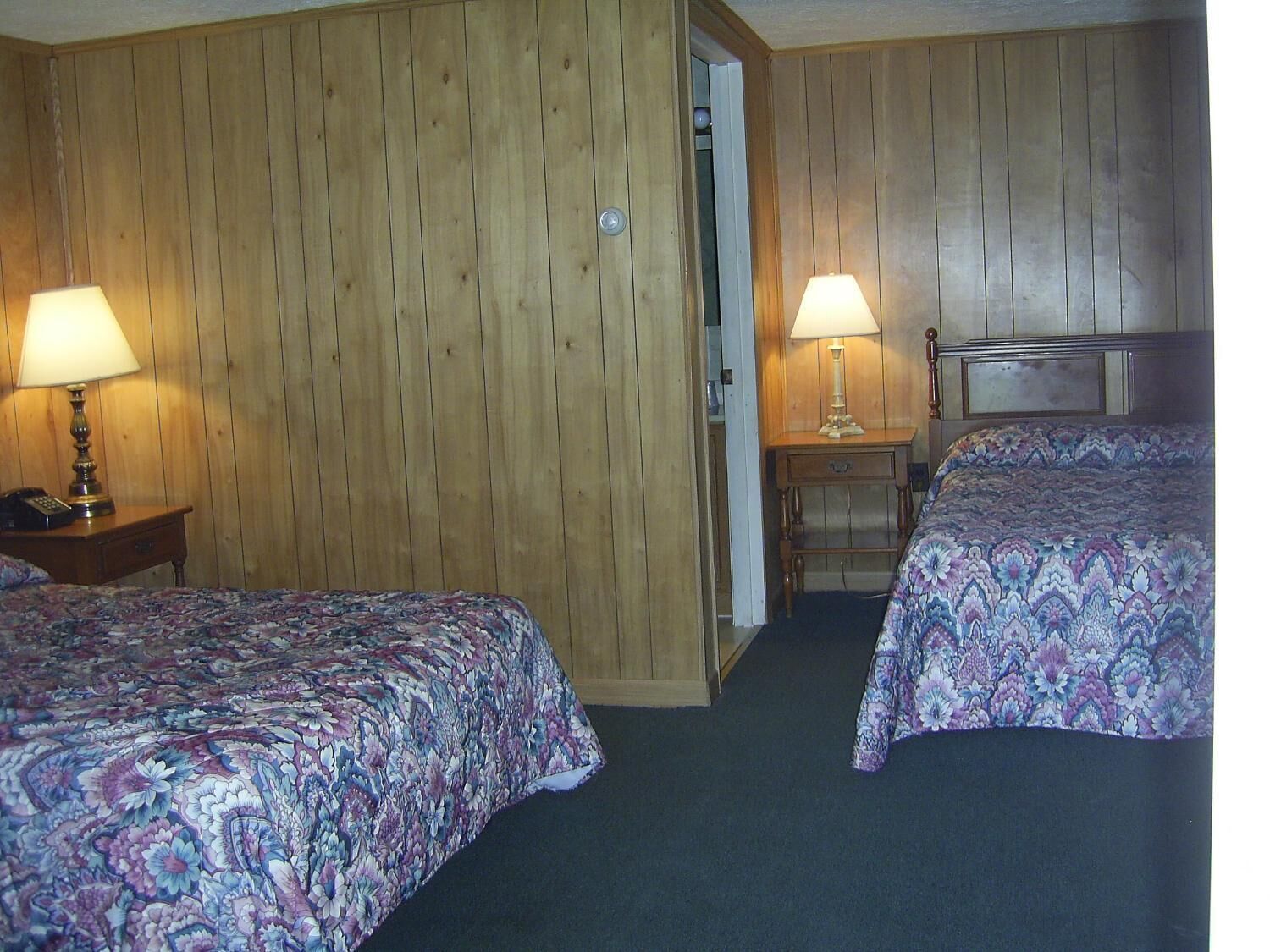Une chambre d’hôtel avec deux lits, des murs en bois, une petite table avec une lampe et une porte donnant sur une autre pièce.