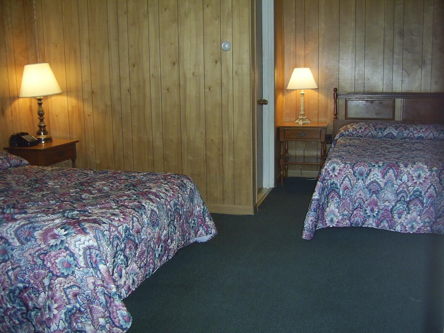 A room with two beds, each with a floral-patterned bedspread, a wooden door, and a small bedside table with a lamp.