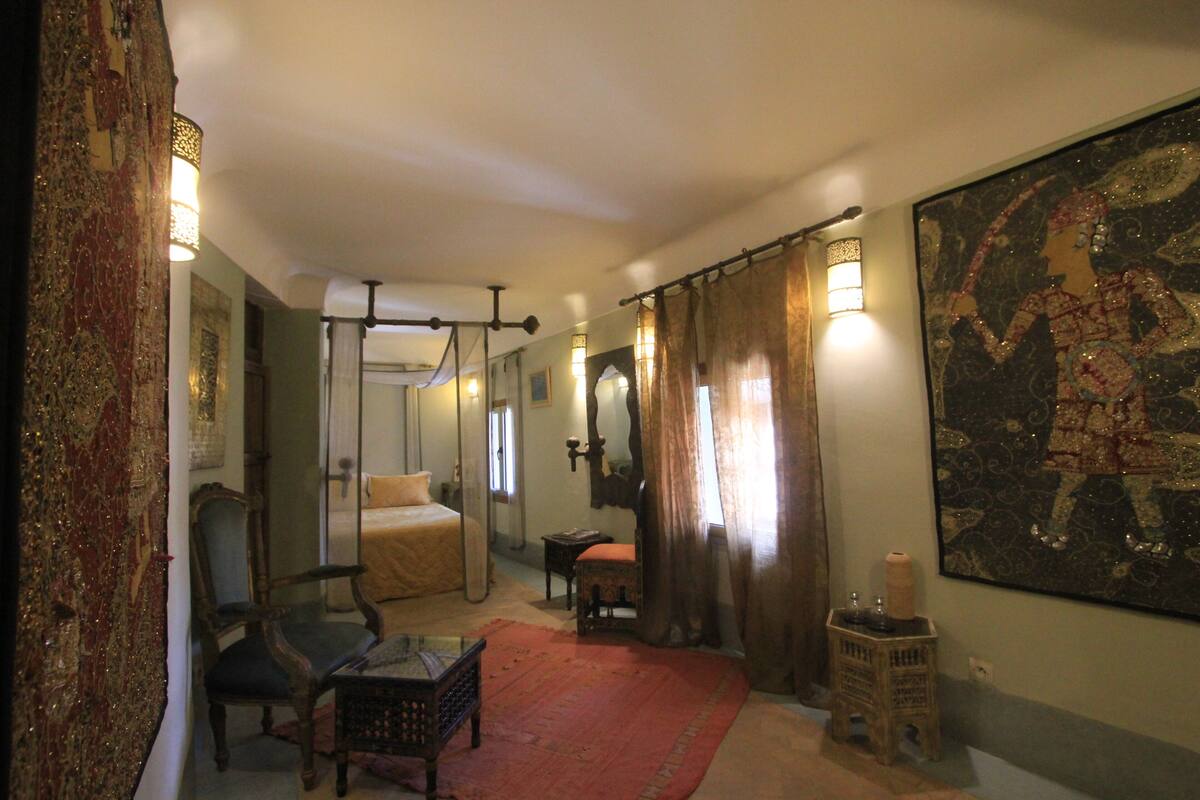 A bedroom with a bed, a chair, a small table, a red rug, and a large tapestry on the wall.