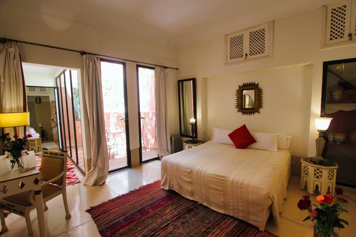 A hotel room with a bed, a desk, a chair, a table with flowers, a lamp, and a view through the glass door.